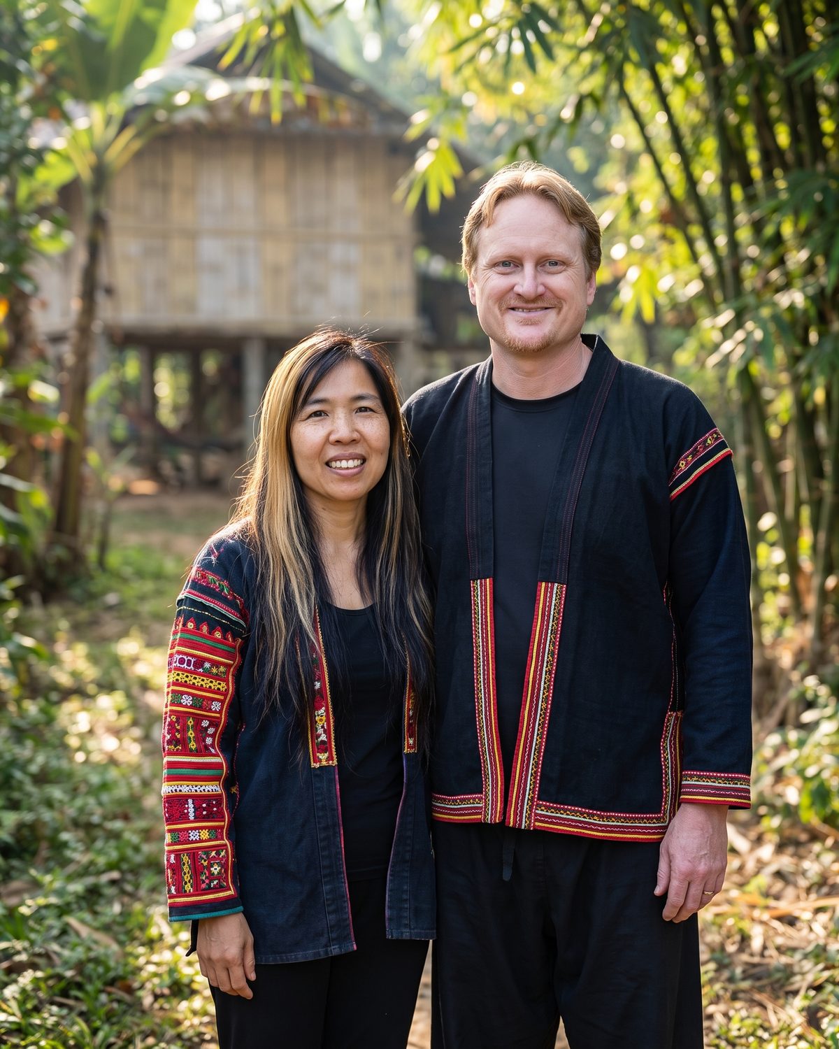 Illustration: Aaron and Alina Glassburn in embroidered Akha jackets in a jungle clearing, a bamboo stilt house behind them.