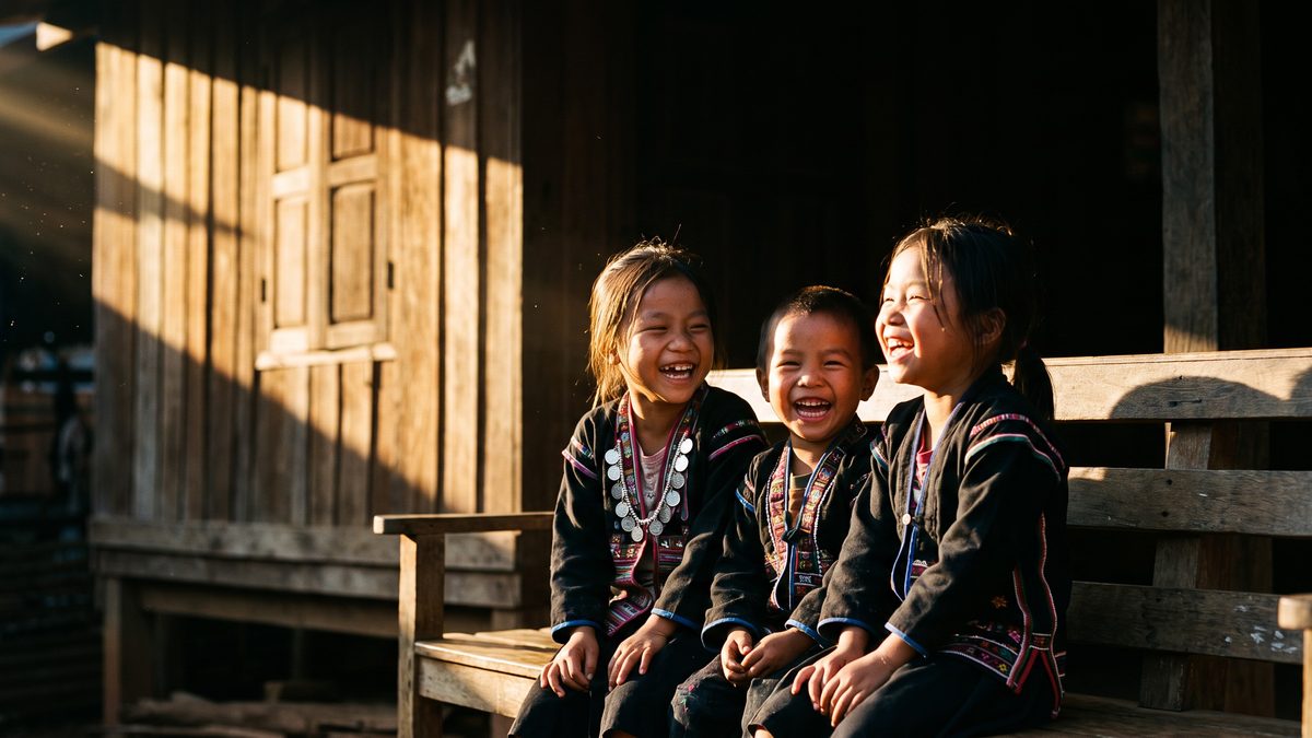 Three hill tribe children in embroidered jackets laugh together on a wooden bench outside a village church in golden afternoon light