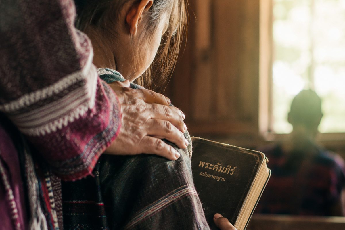 An elder's hand rests on a child's shoulder as the child holds a Thai Bible inside a wooden village church